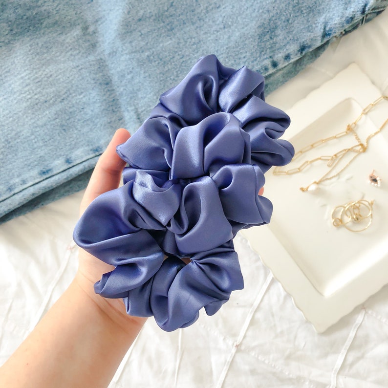 May include: Two blue satin scrunchies, made of soft, silky fabric. The scrunchies are in a hand, and are ready to be used for styling hair.