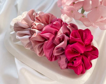 May include: A set of three pink and red satin scrunchies. The scrunchies are arranged in a gradient from light pink to dark red.
