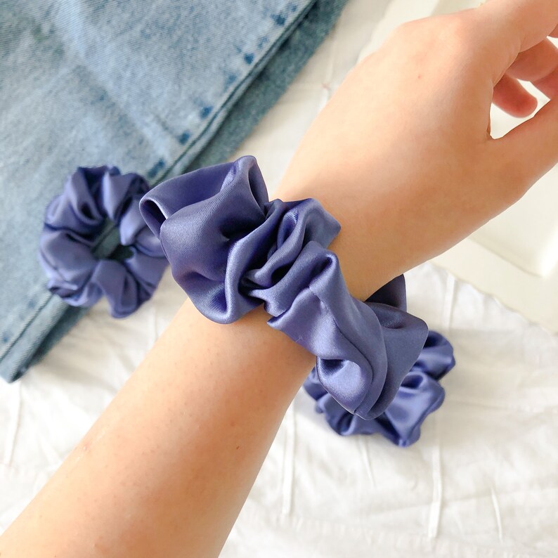 May include: Two purple satin scrunchies, one worn on a wrist and one lying on a white surface.