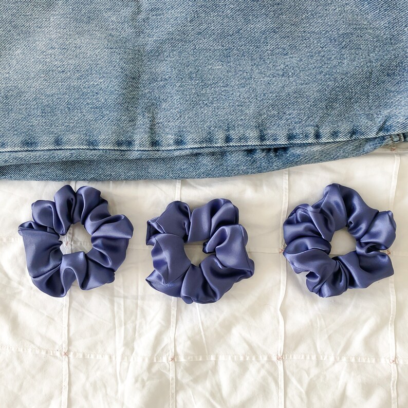 May include: Three purple scrunchies made of soft fabric. The scrunchies are arranged in a row on a white surface.