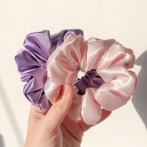 May include: Two satin scrunchies, one purple and one pink, are tied together in a knot. The scrunchies are made of soft, silky fabric and are perfect for holding hair in place.