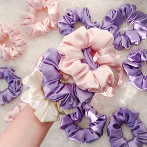 May include: A collection of colorful satin scrunchies in shades of pink, purple, and white. The scrunchies are arranged in a variety of styles and sizes, and they are perfect for adding a touch of color and style to any hairstyle.