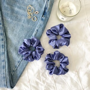 May include: Three purple satin scrunchies on a white surface. The scrunchies are made of soft, silky fabric and are perfect for holding hair in place.