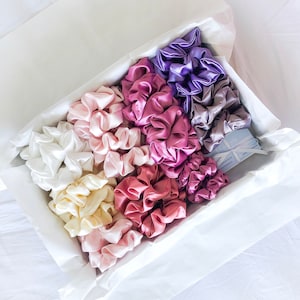 May include: A collection of 12 colorful satin scrunchies in a white gift box. The scrunchies are arranged in a rainbow pattern, with shades of purple, pink, and cream.