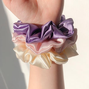 May include: A set of three satin scrunchies in shades of purple, pink, and cream. The scrunchies are layered on top of each other, with the purple scrunchie on top.