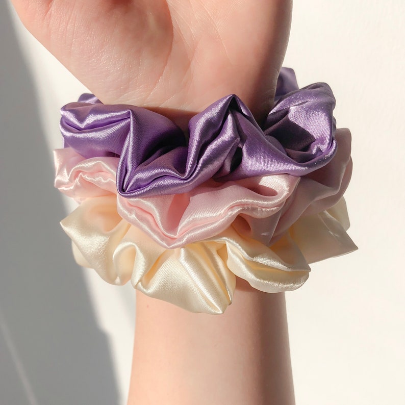 May include: A set of three satin scrunchies in shades of purple, pink, and cream. The scrunchies are layered on top of each other, with the purple scrunchie on top.