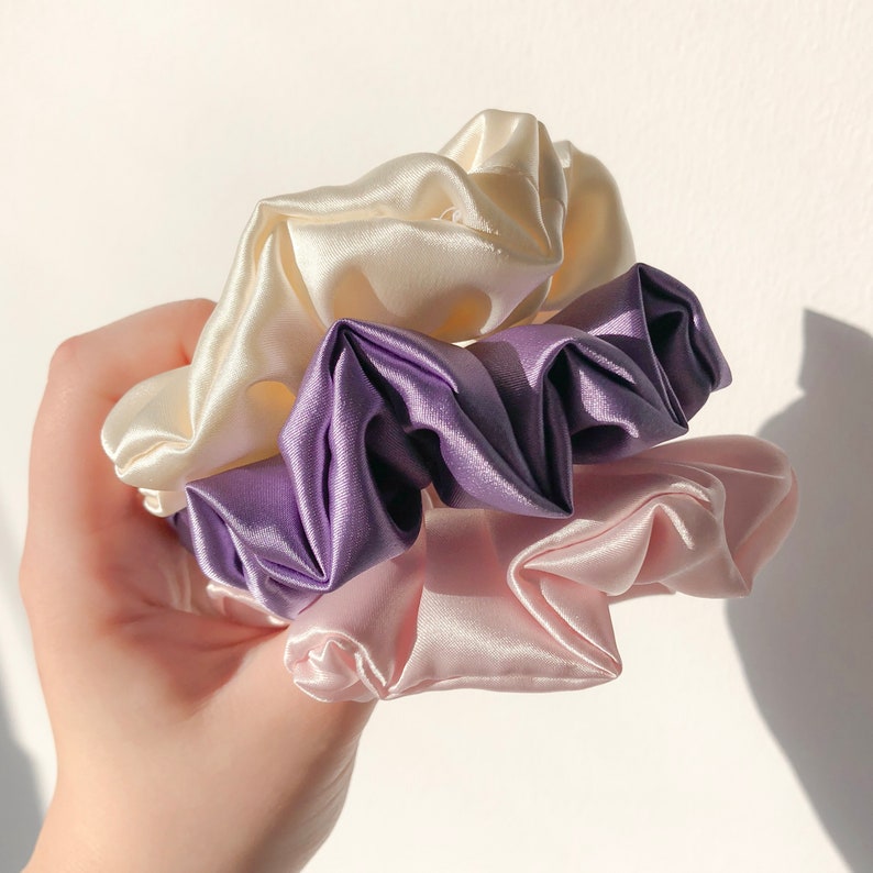 May include: Three satin scrunchies in a stack, the top one is ivory, the middle one is purple, and the bottom one is pink.