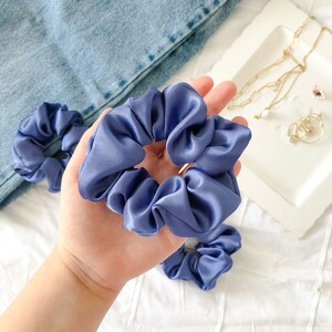 May include: Two blue satin scrunchies, one held in a hand and the other lying on a white surface.