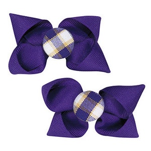 May include: Two hair bows made of deep purple grosgrain ribbon, each featuring a central circular button covered in a white, purple, and gold plaid fabric pattern. The bows have a structured, multi-layered design with pointed ends.