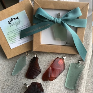 May include: Five sea glass pendants with silver bails, displayed on a light brown fabric surface. The pendants are in various shapes and colors, including green, brown, and red. The text "Seneca Sea Glass" is visible on a box in the background.