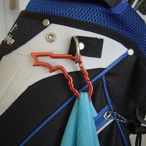 May include: A red metal Florida-shaped carabiner clip attached to a black and blue golf bag. A blue towel is hanging from the clip.