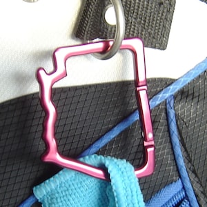 May include: A pink metal carabiner with a unique, textured design. The carabiner is attached to a black and blue bag.