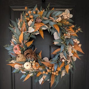 Peut inclure: Une couronne circulaire composée d'un mélange de feuillage vert foncé, de feuilles orange et crème, de petites citrouilles, de pommes de pin et de grappes de baies beige clair. L'arrangement est dense et superposé, créant un aspect texturé.