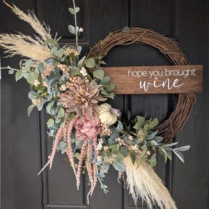 May include: A grapevine wreath decorated with dried pampas grass, dusty green foliage, pink and mauve flowers, and a small white pumpkin. A horizontal wooden sign is attached to the right side with the white text hope you brought wine.