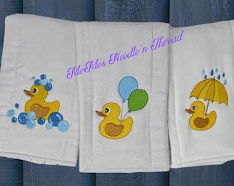 May include: Three white burp cloths with embroidered yellow rubber duckies. The first duck is in a bubble bath, the second is holding balloons, and the third is holding a yellow umbrella in the rain. The text "Me Me's Needle 'n Thread" is embroidered on the top of the middle cloth.