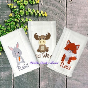May include: Three white burp cloths with embroidered woodland animals and the name "Reid". The animals are a rabbit holding a carrot, a moose, and a fox.