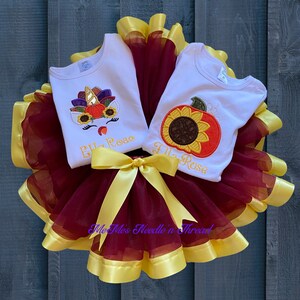 Puede incluir: Dos camisas blancas sin mangas con dise&ntilde;os bordados de tem&aacute;tica oto&ntilde;al. Una camisa presenta un unicornio con una calabaza, girasoles y hojas de oto&ntilde;o. La otra camisa presenta un girasol y una calabaza. Ambas camisas tienen el nombre "Ella Rose" bordado. Las camisas se muestran en una falda de tul marr&oacute;n con una cinta de sat&eacute;n amarilla.