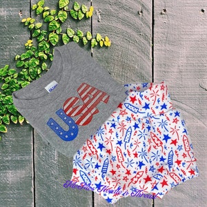 May include: A grey t-shirt with the word "USA" printed in red, white, and blue. The shirt is paired with white shorts with a red, white, and blue firework pattern.