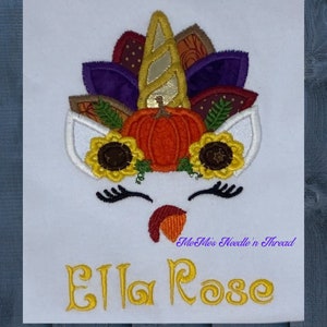May include: Embroidered design of a unicorn with a pumpkin and autumn leaves on its head. The unicorn has a white face with closed eyes and a small, red, turkey-like wattle. The text "Ella Rose" is embroidered below the unicorn. The text "Me Me's Needle n' Thread" is embroidered below the text "Ella Rose".