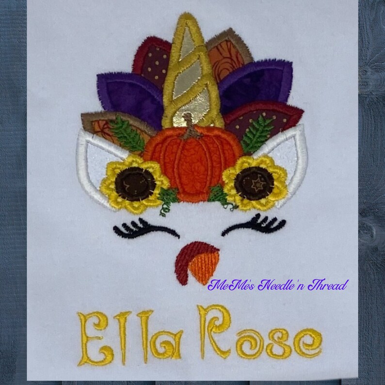 May include: Embroidered design of a unicorn with a pumpkin and autumn leaves on its head. The unicorn has a white face with closed eyes and a small, red, turkey-like wattle. The text "Ella Rose" is embroidered below the unicorn. The text "Me Me's Needle n' Thread" is embroidered below the text "Ella Rose".