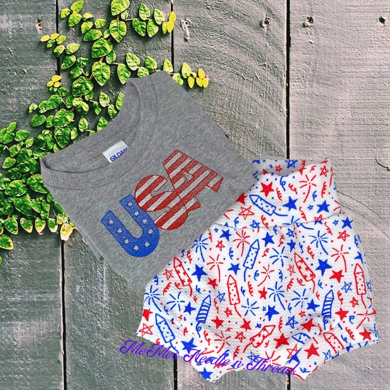 May include: A grey t-shirt with the word "USA" in blue and red letters, with a red, white, and blue American flag design in the "A". The t-shirt is paired with white shorts with a red, white, and blue firework pattern.