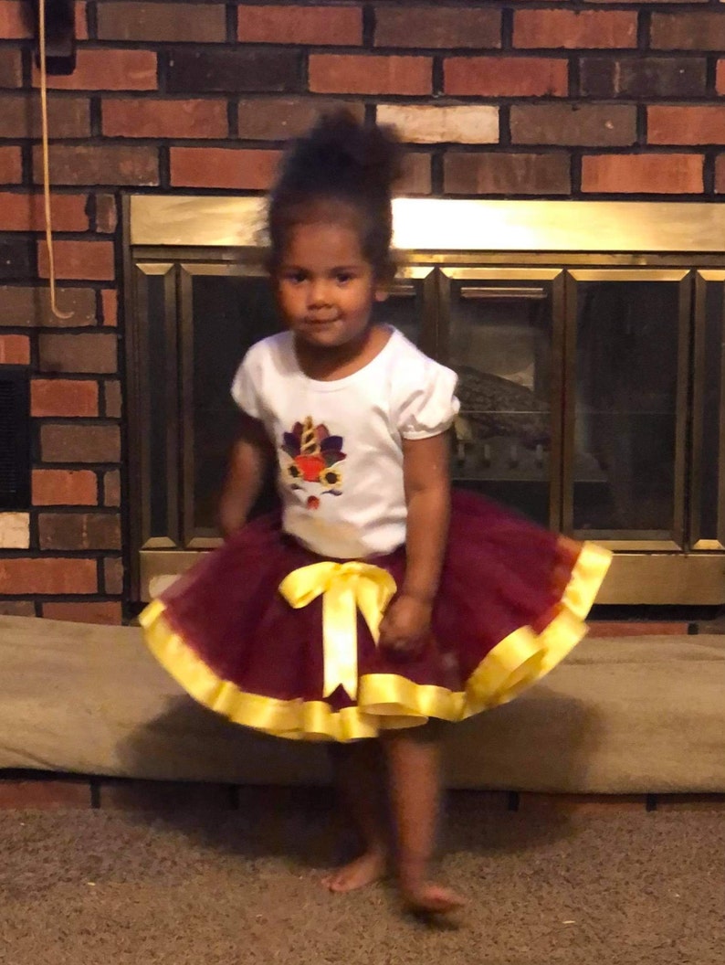 Puede incluir: Una ni&ntilde;a peque&ntilde;a con una camiseta blanca con un dise&ntilde;o de oto&ntilde;o colorido y una falda de tul marr&oacute;n con una cinta de sat&eacute;n amarilla.