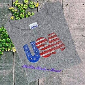 May include: Gray Gildan t-shirt with the word "USA" in red, white, and blue with a glittery finish. The letters are stylized to resemble the American flag. The text "Me Mess Needle 'n Thread" is printed below the word "USA".