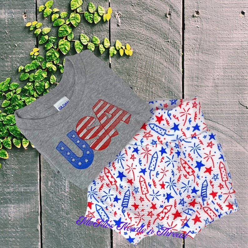 May include: A grey t-shirt with the word "USA" printed in red, white, and blue. The shirt is paired with white shorts with a red, white, and blue firework pattern.