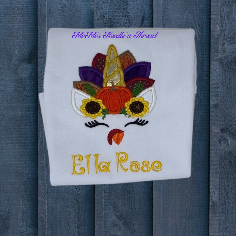 May include: White onesie with an embroidered unicorn design featuring a pumpkin, sunflowers, and autumn leaves. The name "Ella Rose" is embroidered below the unicorn.