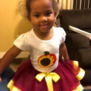 Puede incluir: Una ni&ntilde;a peque&ntilde;a con una camiseta blanca con un dise&ntilde;o bordado de girasol y calabaza. Tambi&eacute;n lleva una falda de tul marr&oacute;n con detalles de cinta amarilla.