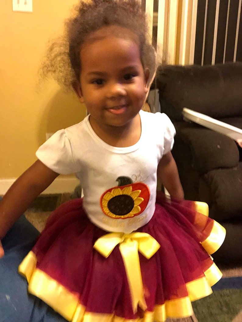 Puede incluir: Una ni&ntilde;a peque&ntilde;a con una camiseta blanca con un dise&ntilde;o bordado de girasol y calabaza. Tambi&eacute;n lleva una falda de tul marr&oacute;n con detalles de cinta amarilla.