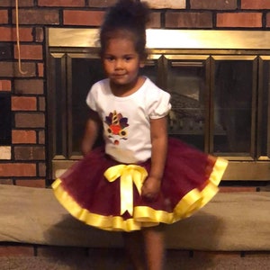 May include: A young girl wearing a white t-shirt with a colourful turkey design and a maroon tulle skirt with a yellow ribbon.
