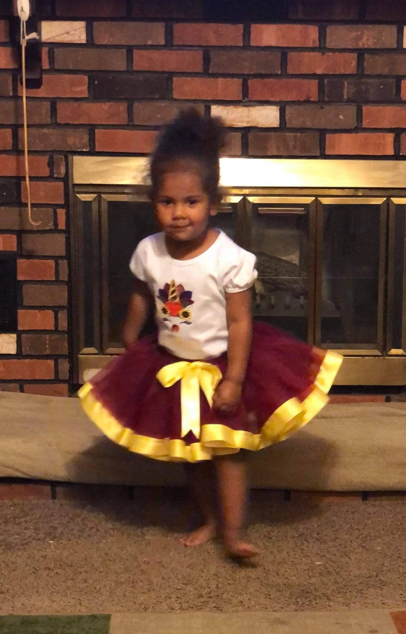 May include: A young girl wearing a white t-shirt with a colourful turkey design and a maroon tulle skirt with a yellow ribbon.