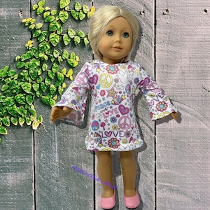 May include: A doll with blonde hair wearing a white long-sleeve dress featuring a colorful pattern of peace signs, hearts, stars, flowers, and the text "Peace and LOVE". The doll has light skin, green eyes, and is wearing solid pink flat shoes.