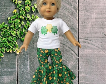 May include: A doll with blonde hair wearing a white short-sleeved t-shirt featuring three embroidered shamrocks in green and yellow. The doll is also wearing flared pants with an all-over print of small green and yellow shamrocks on a dark background.