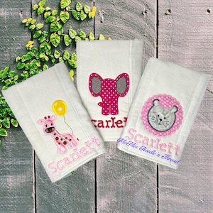 May include: Three white burp cloths with pink and gray embroidered animal designs. The designs include a giraffe, an elephant, and a lion. Each cloth has the name "Scarlett" embroidered on it. The bottom cloth also has the text "The  Alfie's Needle 'n Thread" embroidered on it.