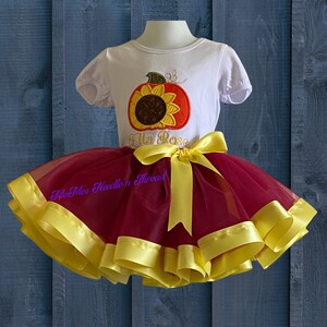 Puede incluir: Una camiseta blanca con una calabaza bordada con una cara de girasol y el texto "Ella Rose". La camiseta se combina con un tut&uacute; de tul marr&oacute;n con ribete de cinta de sat&eacute;n amarilla.