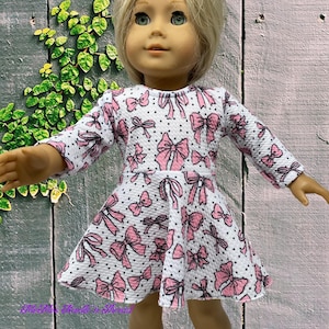 May include: A doll wearing a white dress with a pink bow pattern and black polka dots. The dress has long sleeves and a flared skirt. The doll has blonde hair and green eyes.