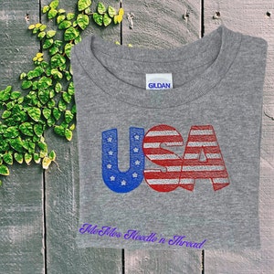 May include: A gray t-shirt with the word "USA" printed in red, white, and blue. The letters are stylized to look like the American flag. The text "Me Mess Needle n Thread" is printed in purple below the word "USA".