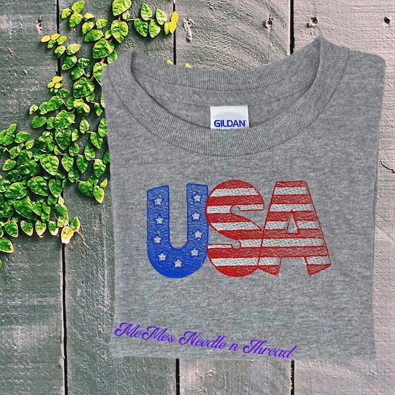 May include: A gray t-shirt with the word "USA" printed in red, white, and blue. The letters are stylized to look like the American flag. The text "Me Mess Needle n Thread" is printed in purple below the word "USA".