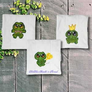 May include: Three white fabric squares with embroidered green frogs. One frog is wearing a gold crown, one is holding a yellow balloon, and one is wearing a pink bow. The squares have the text "Me, Miss Needle 'n Thread" on the bottom.
