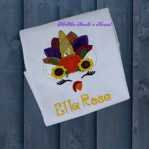 May include: White long-sleeved shirt with an embroidered unicorn design featuring a pumpkin, sunflowers, and autumn leaves. The shirt also has the name "Ella Rose" embroidered in yellow thread.