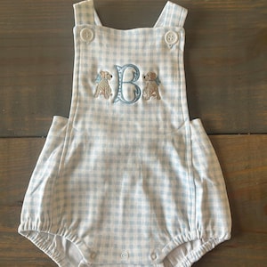 Puede incluir: Un pelele de bebé de gingham azul y blanco con una letra "B" bordada en blanco y dos perros bordados en la parte delantera.