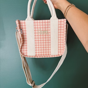 May include: A small, pink and white gingham tote bag with a cream-colored shoulder strap and handles. The word "Flora" is embroidered in a light green thread on the front of the bag. The bag is held up by a person.