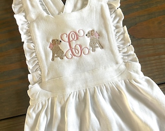 Könnte beinhalten: Weißer Baby-Strampler mit Rüschen am Ausschnitt und Trägern. Der Strampler hat ein Monogramm "C" mit zwei gestickten Hunden, die rosa Schleifen tragen.