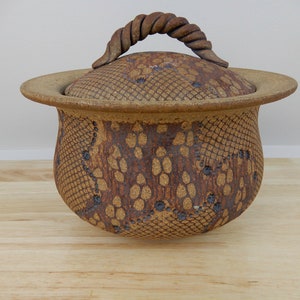 May include: A ceramic lidded jar with a textured surface and a twisted handle. The jar is primarily tan with brown and dark blue accents. The lid has a wide brim, and the body of the jar has a patterned design.