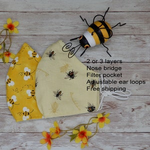May include: A reusable face mask with a yellow and white bee pattern. The mask has a nose bridge, filter pocket, and adjustable ear loops. The text "2 or 3 layers, Nose bridge, Filter pocket, Adjustable ear loops, Free shipping" is written on the mask.