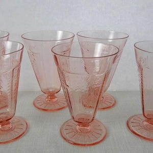 May include: A set of five pink glass tumblers with a footed base and a textured, embossed pattern on the exterior. Each glass features a flared rim, a tapered body, and a circular base with radiating lines, creating a uniform appearance for the collection.