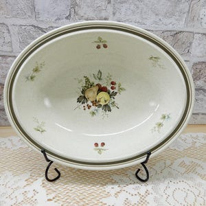 May include: An oval ceramic serving bowl with a speckled cream finish and a dark brown rim. The interior features a central botanical illustration of a pear, apple, and berries, surrounded by smaller sprigs of fruit and leaves around the edge.