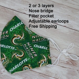 May include: A green face mask with a repeating pattern of the words "CHARLOTTE" and "NINER NATION" in white and gold. The mask has adjustable earloops and a filter pocket. The text "2 or 3 layers, Nose bridge, Adjustable earloops" is also visible.